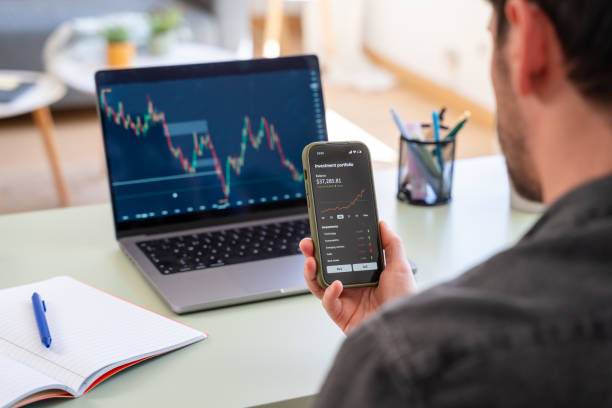 Rear view of a man checking investment portfolio on mobile phone with stock market graphs displayed on a laptop screen
